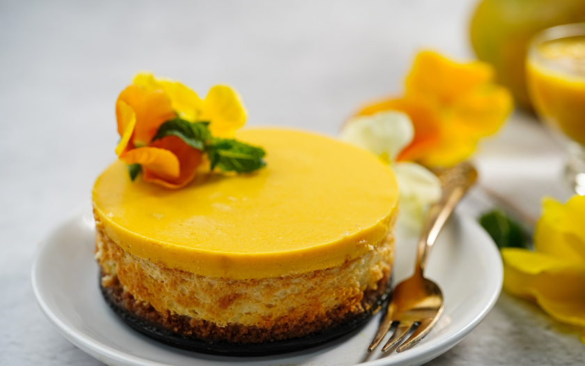Chiếc bánh Mousse vàng tươi rực rỡ, bề mặt láng mịn, được trang trí bằng hoa tươi và lá bạc hà